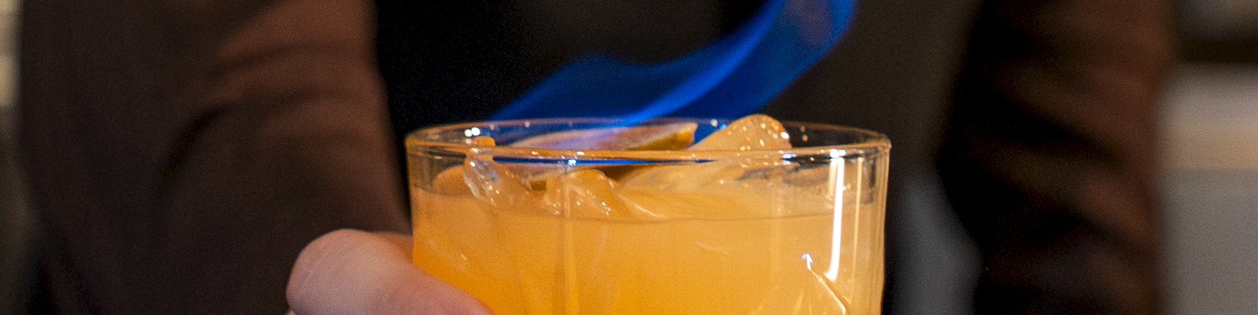 A bartender holds a glass of orange cocktail with a blue flame rising from the top.