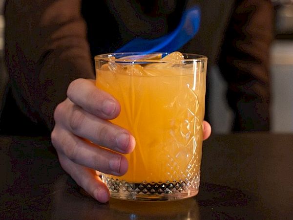 A bartender holds an orange cocktail in a crystal glass, with a blue flame/smoke effect above it.