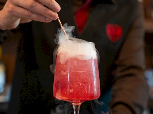 A bartender pours a smoky red cocktail into a glass using a copper straw.