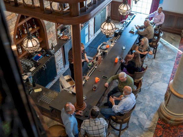 A group of people gathers at a bar with a bartender, drinks and glasses on the counter in a warmly lit lounge.