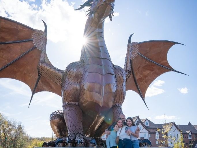 A large dragon-shaped statue rises behind three people posing in front of it under a bright blue sky.