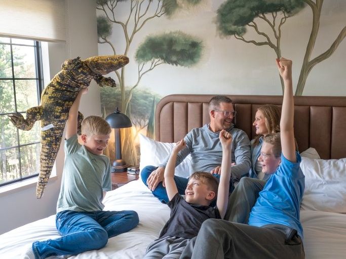 A family plays in a bedroom with a plush dinosaur in front of a tree mural while kids cheer and jump on the bed.