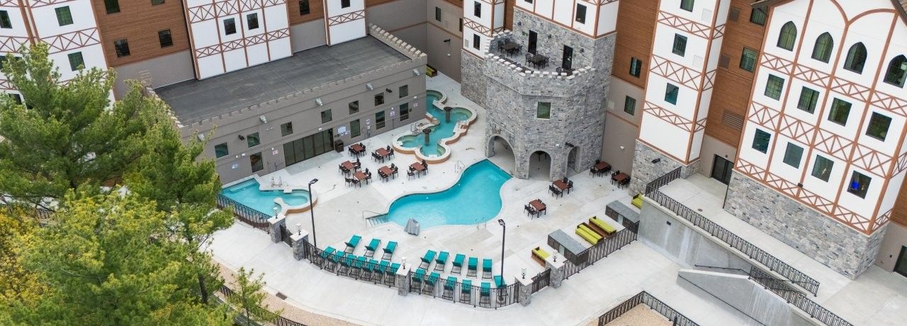 A medieval-style courtyard hotel with white-and-brown timber buildings, an outdoor pool, lounge seating, and a stone archway.