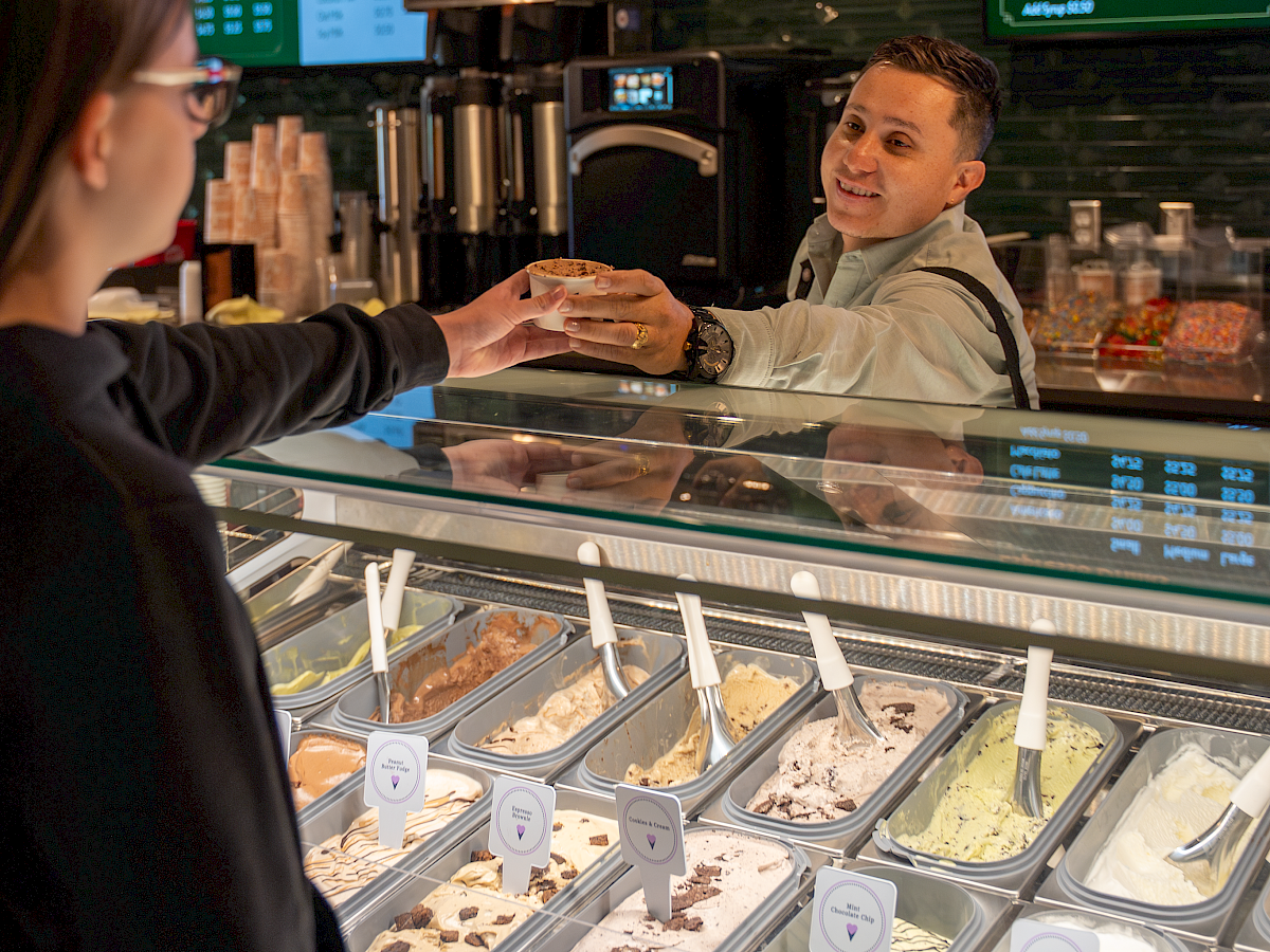 A barista hands a customer a cup of coffee/espresso in a coffee shop, with an ice-cream display and menu boards behind them.