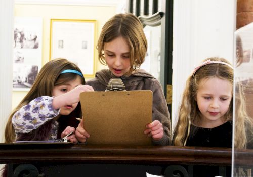 Three kids and an adult huddle over a cardboard folder on a table, collaborating and looking closely together.