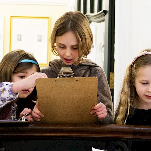 Three kids and an adult huddle over a cardboard folder on a table, collaborating and looking closely together.