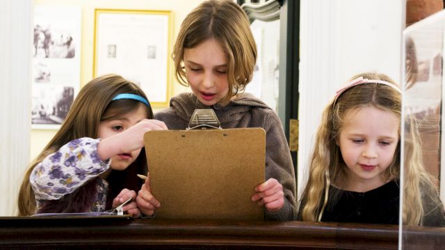 Three kids and an adult huddle over a cardboard folder on a table, collaborating and looking closely together.