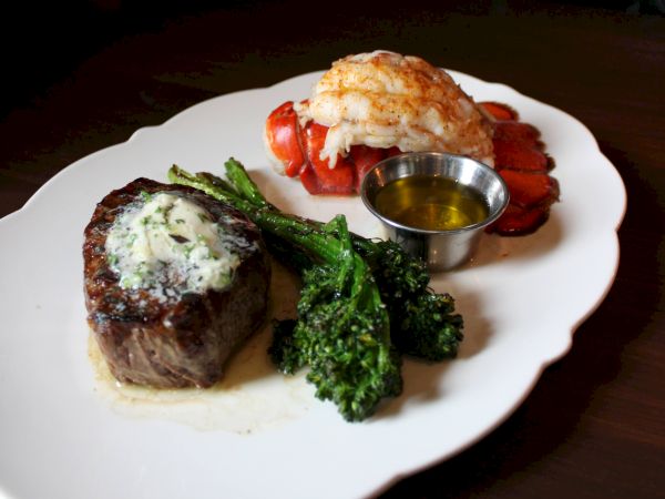 A plated steak with lobster/shrimp, saut&eacute;ed broccolini, and a small cup of butter sauce on a white dish.
