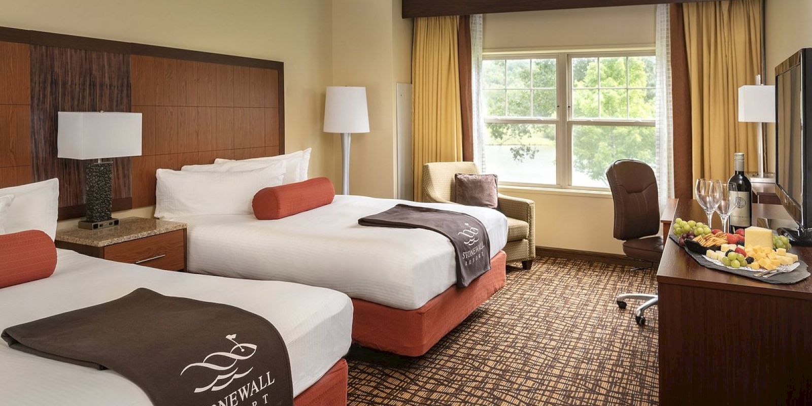 The image shows a hotel room with two beds, a window, a TV, chairs, and a table with wine and snacks on it.