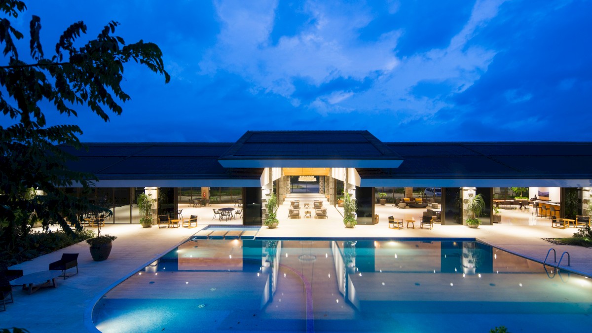 A modern building with open interiors and a lit swimming pool, surrounded by trees, under a deep blue evening sky.