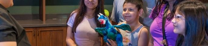 A family smiles at a store counter as a child holds a colorful stuffed animal and a clerk rings up the purchase.
