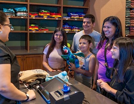 A family smiles at a store counter as a child holds a colorful stuffed animal and a clerk rings up the purchase.