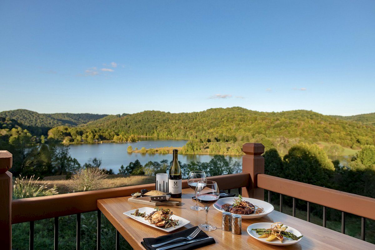 A scenic outdoor dining setup with wine, plates of food, and a view of a lake and lush green hills under a clear blue sky.