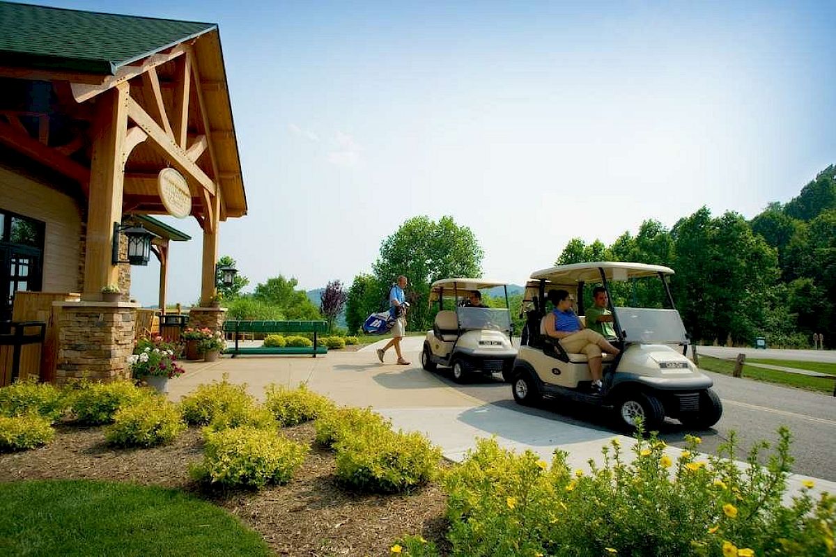 Golfers with bags and people in golf carts are outside a clubhouse, surrounded by greenery on a sunny day.