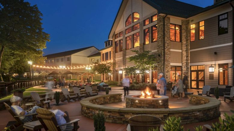 The image shows a cozy outdoor area at night with a lit fire pit, surrounded by chairs, a building, and string lights.