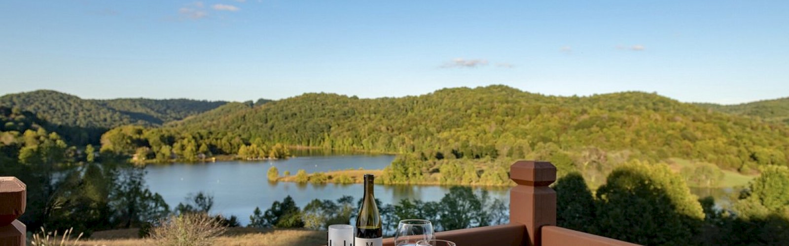 A scenic view of a lake and hills from a balcony, with a table set for dinner including wine, plates of food, and glasses.
