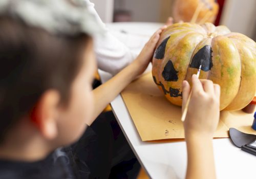 Kids paint a large pumpkin on a table with brushes, creating a spooky face for a Halloween decoration.