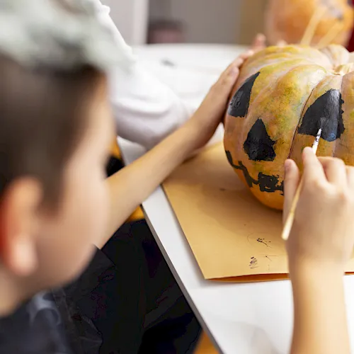 Kids paint a large pumpkin on a table with brushes, creating a spooky face for a Halloween decoration.