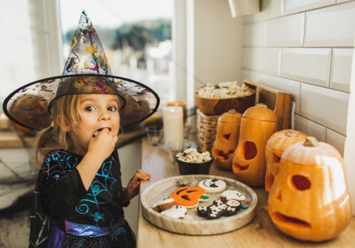 A little girl in a Halloween witch costume surprises a cookie while sitting at a table with jack-o&rsquo;-lanterns and treats in the kitchen.