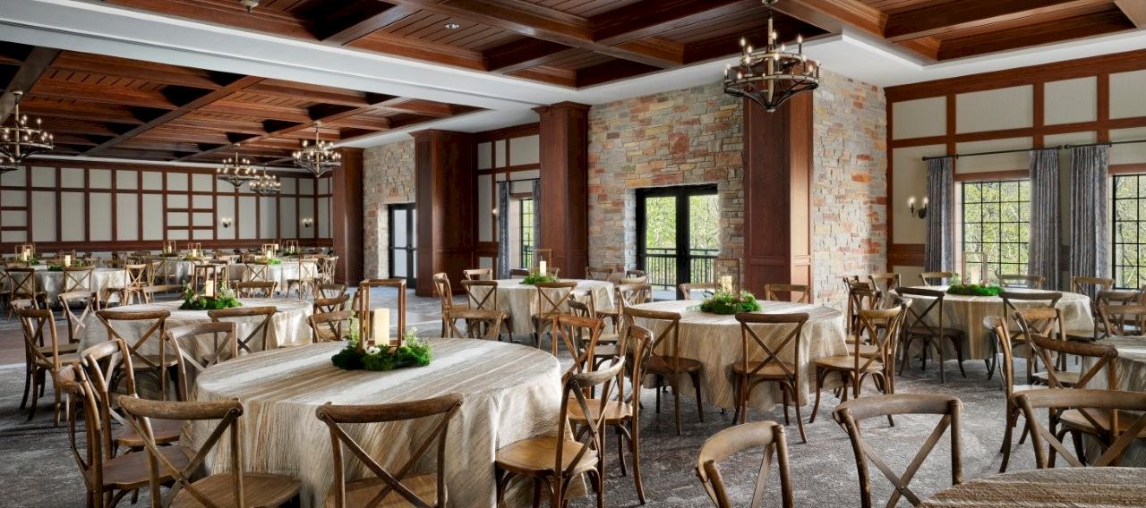A rustic banquet hall with round wooden tables, linen-covered chairs, stone walls, and ornate chandeliers under dark wooden beams.