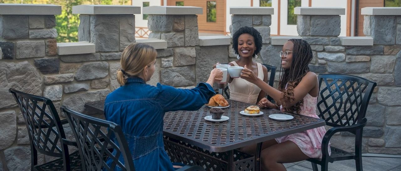 Three friends sit at an outdoor table, chatting and sharing pastries in front of a stone wall and building.
