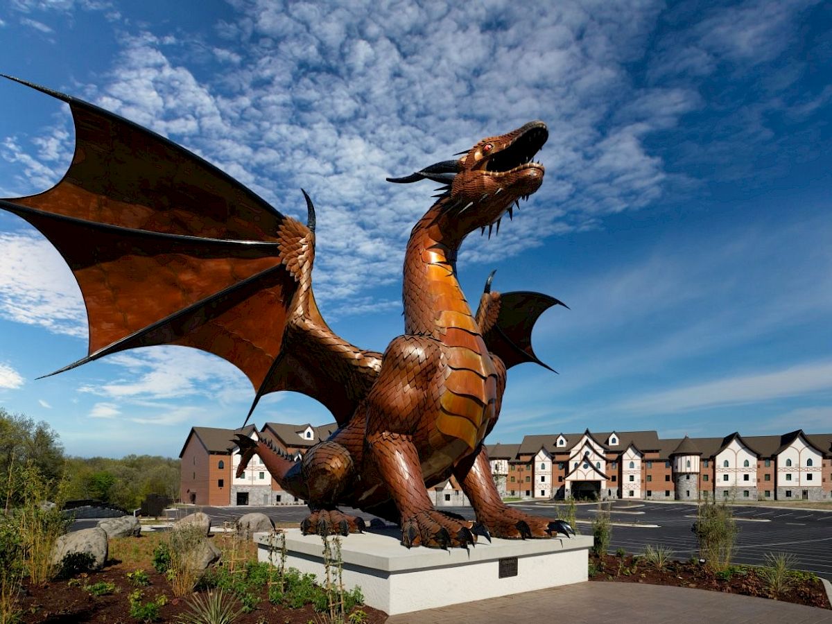 A bronze dragon sculpture with wings spread stands on a pedestal in front of buildings under a blue sky with clouds.