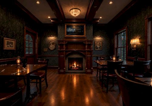 A cozy dining room with a lit fireplace, dark wood mantel, chandelier overhead, and set tables by large windows.