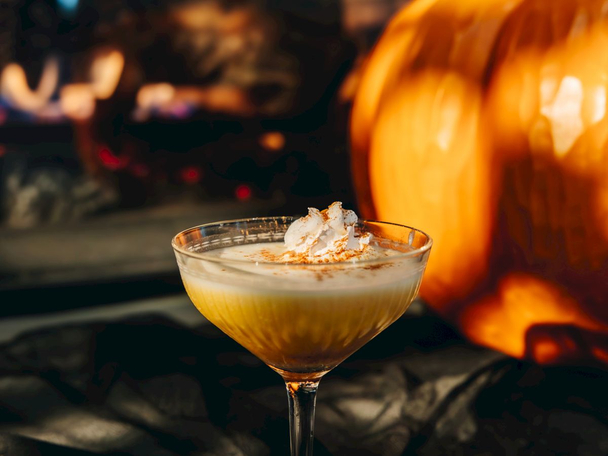 A cocktail glass with ice and an amber drink sits by the fire in a bar, with a blurred crowd in the background.