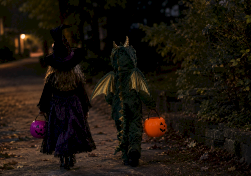 Two kids in Halloween costumes walk at night, holding jack-o&rsquo;-lantern pumpkins&mdash;witch hat and a green unicorn outfit leading the trick-or-treat.