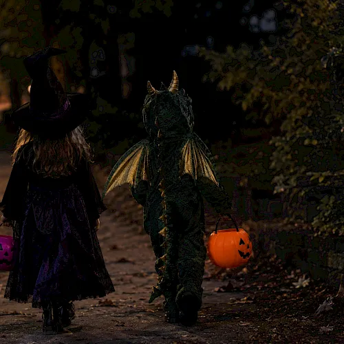 Two kids in Halloween costumes walk at night, holding jack-o&rsquo;-lantern pumpkins&mdash;witch hat and a green unicorn outfit leading the trick-or-treat.