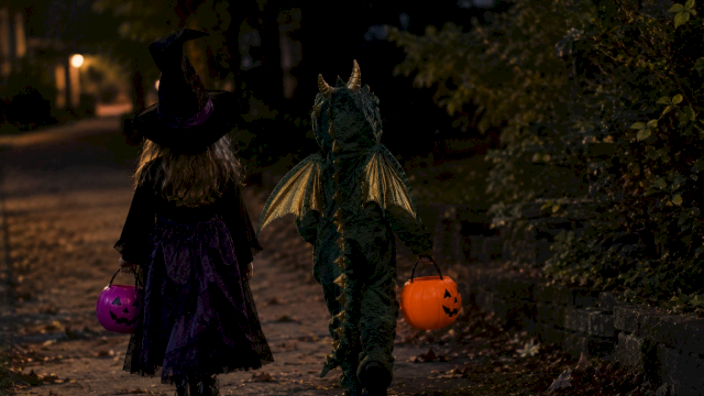 Two kids in Halloween costumes walk at night, holding jack-o&rsquo;-lantern pumpkins&mdash;witch hat and a green unicorn outfit leading the trick-or-treat.