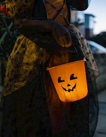 A person in spooky Halloween costume carries an orange jack-o&rsquo;-lantern trick-or-treat bucket with black candy symbols.